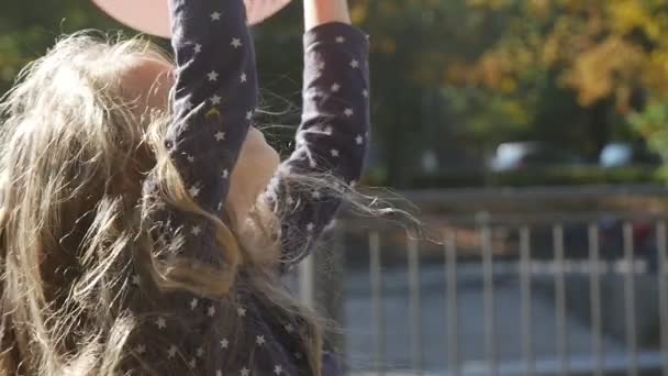 긴 공정한 머리를 가진 어린 소녀는 공을 던져 공을 잡는다 공을 잡는다 점프 공원 나무 울타리 강 배경 화창한 날에 — 비디오