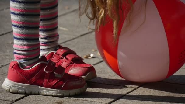 Petite fille aux longs poils blonds joue aux jambes de balle dans des baskets rouges Fermer Fille sourit Saut Parc Arbres Clôture sur fond Journée ensoleillée — Video