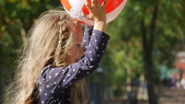 Petite fille avec de longs cheveux blonds joue à la balle lance la balle attrape la balle fille sourit sauter parc arbres clôture sur fond ensoleillé jour — Video