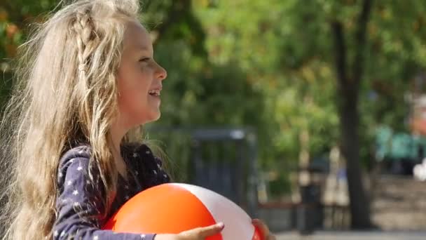 Маленька дівчинка особою крупним планом з довгими справедливими волоссям грає м'яч проведення м'яч дівчина посміхається парк дерева паркан на фоні сонячний день — стокове відео