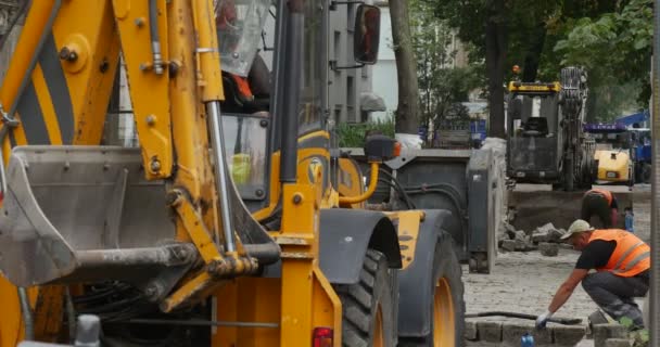 Trabajadores en ropa de trabajo naranja en la excavadora amarilla de reparación de carreteras Close Up Backside Scoop Close Up Pavimentación de la carretera con bloques de árboles a lo largo de la calle de la ciudad — Vídeos de Stock