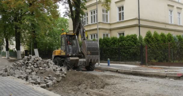 Pilote en tenue de travail Excavatrice jaune saisit les blocs Réparation de la route Pavage de la route avec des blocs La femme de bâtiment marche sur les arbres roulés avec Polietilen — Video