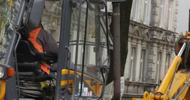 Bestuurder man in oranje werkkleding in cabine van gele graafmachine machine stands Scoop Road Repair werknemers zijn het bestrating van de weg met blokken gebouwen stad — Stockvideo