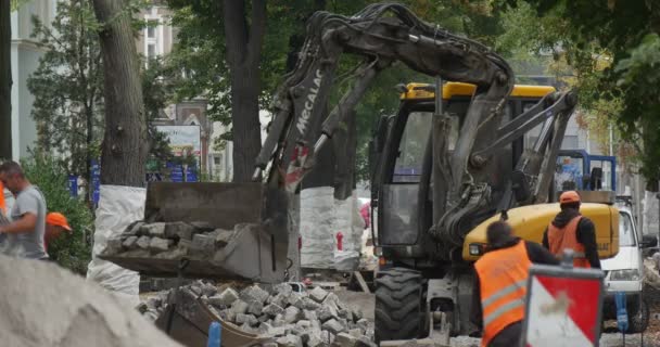 Workers in Orange Workwear Are Working at Road Repair Yellow Excavator with Flasher Driver Scoop is Loading the Blocks Paving the Road with Blocks Trees — Stock videók