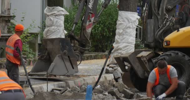 Pracovníci ve žlutých pracovních oděvech převážejí bloky kamenů na ulici Road opravy žluté bagr se pohybuje jeho Scoop Close Up dláždění silnice — Stock video