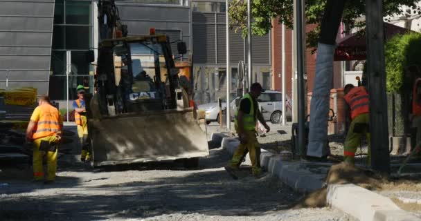 Arbetare i gula arbetskläder på Street Road Repair Yellow Excavator CAT rör sig mot Camera Street Bussar passerar bakom grävmaskinen — Stockvideo