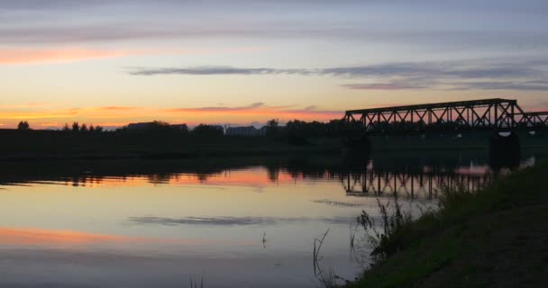 Přichází muž v bílém, stojící na řeky břehu řeky rybník hladké vody stromy siluety reflexe Bridge přes řeku růžové a žluté nebe — Stock video