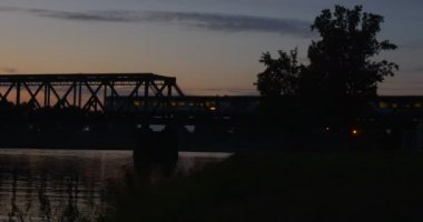 Tren Köprüsü nehir su birikintisi Köprüsü aracılığıyla nehir dalgalanan su ağaçlar siluetleri yansıma Bright günbatımı pembe ve sarı gökyüzü hareketli olduğunu