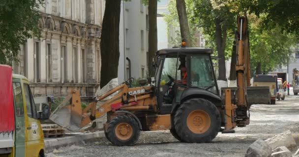 Bestuurder binnenkant van cabine van gele graafmachine staande gebouwen werknemers in workwear op de straat weg reparatie bestrating van de wegen bomen — Stockvideo