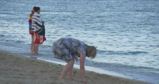 Две девушки, завернутые в полотенца, стоят в воде на мелководье, у моря, женщина идет, люди отдыхают на песчаном пляже, на берегу моря — стоковое видео