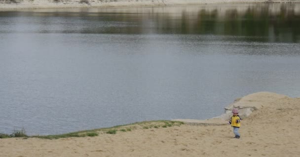 Little Girl Child in Yellow Clothes is Standing at The River Lake Granddaughter is Walking and Falling at Sandy Bank Outdoors Cloudy Green Trees — Stock Video