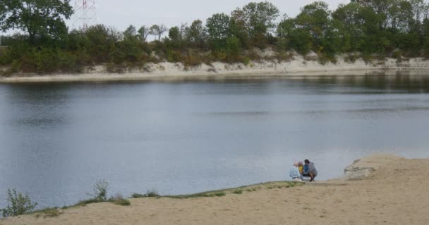 Familia en el lago del río Hombres y niños Padre Abuelo Nieta están sentados en el banco de arena Rippling agua al aire libre Nublado árboles verdes — Vídeos de Stock