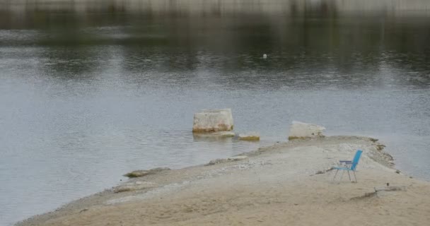 Blauer Stuhl steht am sandigen Ufer Halbinsel plätscherndes Wasser im Freien trübe grüne Bäume — Stockvideo
