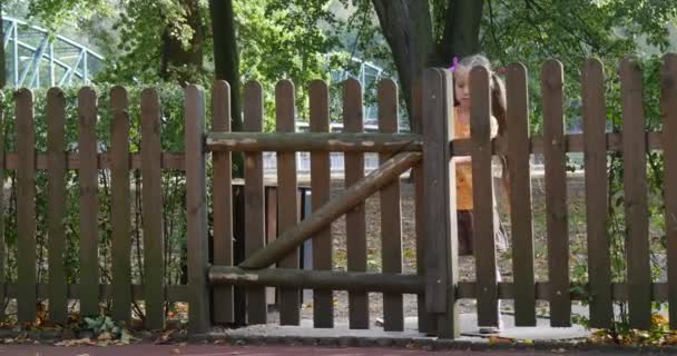 Mädchen versucht, die Tür in Holzzaun Eingang zum Hof zu öffnen Kleines Mädchen mit langen blonden Haaren in orangefarbenem T-Shirt spielt auf Spielplatz sonnig — Stockvideo