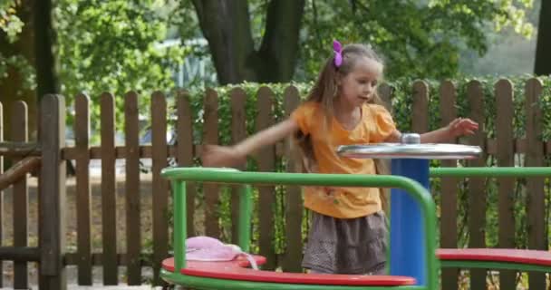 Menina está girando o coelho de brinquedo rosa carrossel no homem carrossel vem menina com longos cabelos justos em laranja t-shirt está jogando s no parque infantil — Vídeo de Stock