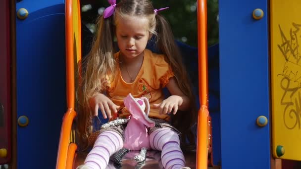 Ragazza è seduta sullo scivolo con il giocattolo rosa Tilda Rabbit Girl sta scendendo bambina con lunghi capelli biondi in arancione T-shirt sta giocando parco giochi — Video Stock