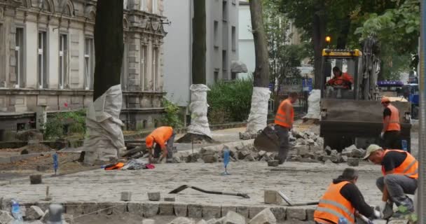 Narancssárga munkaruha az úton dolgozók javítás sárga kotró zár ki hátul összelapátol közel kikövezve az út blokkok fák mentén a város utca — Stock videók