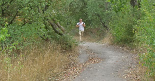 Man τουρίστας άνθρωπος με λευκό μπλουζάκι με σακίδιο είναι τρέχει πάνω από το μονοπάτι στο πράσινο λόφο προσεγγίσεις στην κάμερα κατάφυτη πράσινο λόφο πράσινα δέντρα — Αρχείο Βίντεο