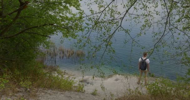 Mann Tourist Mann in weißem T-Shirt Mann mit Rucksack steigt ins Wasser nahm seinen Rucksack ab sitzt am Wasser unter grünem Hügel — Stockvideo