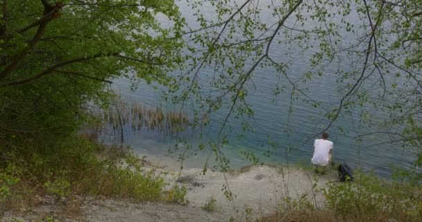 Man Tourist Man in White T-Shirt Man With Backpack is Sitting at the Water Man Gets Up Have Took His Backpack On and Climbing Up by the Green Hill — Stok Video