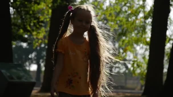 Το κορίτσι είναι στροφή γύρω από το ήλιο χαμογελώντας αργή κίνηση λίγο κορίτσι με μακρά δίκαιη τρίχες δύο αλογοουρές λιλά τόξα πορτοκαλί T-Shirt κορίτσι στην παιδική χαρά — Αρχείο Βίντεο
