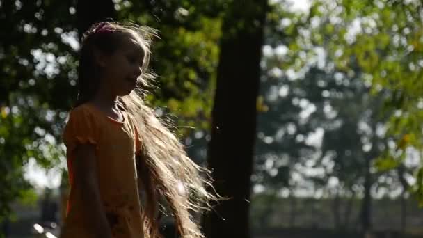 Mädchen dreht sich um in der Sonne lächelnd Zeitlupe kleines Mädchen mit langen blonden Haaren zwei Pferdeschwänze lila Schleifen orange T-Shirt Mädchen auf dem Spielplatz — Stockvideo