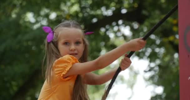 Mädchen mit langen blonden Haaren klettert b auf die Rutsche, hält am Seil und lächelt hinunter, kleines Mädchen mit langen Haaren Pferdeschwanz lila Schleifen T-Shirt Kinder — Stockvideo