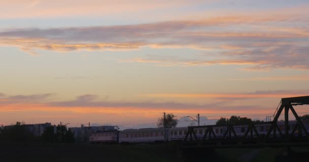 旅客列車は都市を通して、川川銀行の建物反対銀行夕焼け夕日青い空ピンク雲に向かって橋で移動 — ストック動画