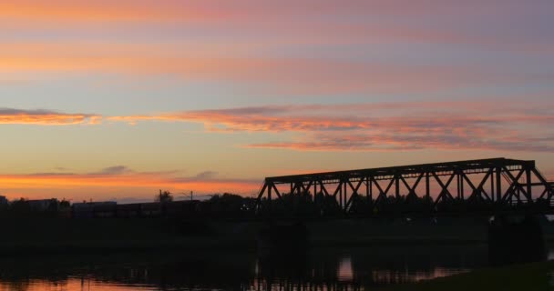 Tren de carga Distante orilla del río se mueve por puente a través de los edificios del río Crepúsculo de la ciudad Nubes son luz al atardecer Reflejo del cielo en el agua — Vídeos de Stock