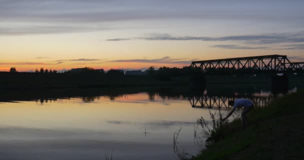 Mann in Weiß kommt ans Flussufer Mann spricht mit Handy Fluss Teich plätschernde Wasserbäume Silhouetten Spiegelbrücke rosa und gelb Himmel — Stockvideo