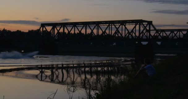 Beyaz adam River banka su Bisiklet döner nehir su birikintisi dalgalanan su ağaçlar siluetleri yansıma köprü günbatımı pembe ve sarı gökyüzü üzerinde oturuyor — Stok video