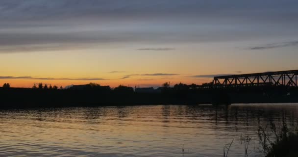 Le train se déplace par le pont Rivière Les gens sur un vélo sur un horizon Ondulation Silhouettes des arbres aquatiques Reflet Coucher de soleil lumineux Ciel rose et jaune — Video