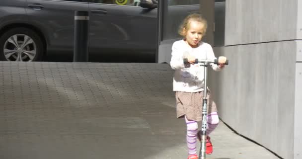 Meisje op een Kick Scooter benaderingen de Camera meisje in rok met Blonde haren is Smiling bevrijden van een Kick Scooter op de straat lopen mensen auto 's — Stockvideo