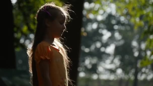 La fille se retourne au soleil Sourire Petite fille au mouvement lent avec de longs cheveux blonds Deux queues de cheval Lilas arcs T-shirt orange fille au terrain de jeux — Video