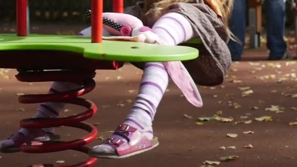 Девушка качается на качелях со своим игрушечным кроликом Розовая Тильда качается на качелях Маленькая девочка с длинными светлыми волосами в оранжевой футболке на детской площадке — стоковое видео