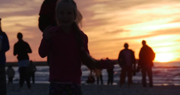 Маленька дівчинка в рожевому з тасьмою Strews пісок повороту біжить люди силуети ходьба піщаний пляж жовтий захід кайт фестиваль Леба Польща — стокове відео
