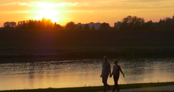 Para mężczyzna i kobieta w ciąży kobieta sylwetki są spacery wzdłuż rzeki Sunset River Bank wody przepływ budynków na przeciwnym brzegu drzew sylwetki — Wideo stockowe