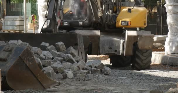 Gele graafmachine grijpt de Heap van blokken en hen vervoert in Scoop Heap van blokken een andere graafmachine Scoop op voorgrond Street weg reparatie zonnige — Stockvideo