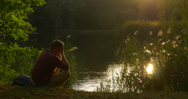 Человек сидит на берегу озера Силуэт человек держит несколько лезвий Положите их вниз Покрыть лицо руками солнце светит к лицу Маллард плавает — стоковое видео