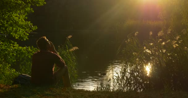 Muž sedí u siluety jezerní banky muž má na sobě svůj obličej s ručičky slunce k jeho obličeji divoká kachna je Plaveck přerostlá banka
