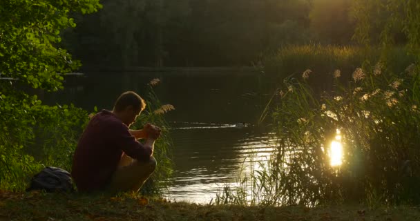 Человек сидит на берегу озера со сложенными руками, склоненная голова Человек молится рюкзак близко к человеку, репеллинг воды Отражение солнца в воде — стоковое видео