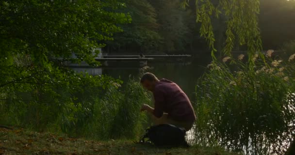 男は湖の銀行の男に座っている彼のバックパックを取った男は横たわって、彼の頭の下にバックパックを置く緑の枝を保持しています — ストック動画