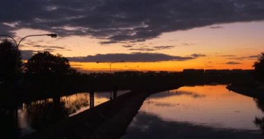 City Dike'deki Nehir Barajı Barajı, Pürüzsüz Su Yansıması Residental Houses Distantly Bright Yellow Sunset Trees'in Siluetlerini Destekliyor