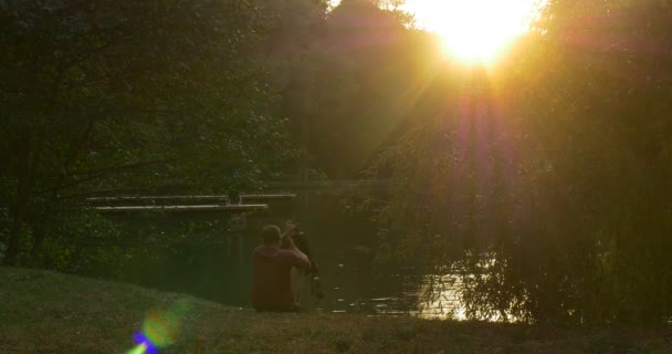 Man is lying at The Lake Bank on The Grass Man Has Stood Up Took the Backpack On and Walking Away Sun Shines to the Camera Sun Behind the Trees Sunset — Vídeo de Stock