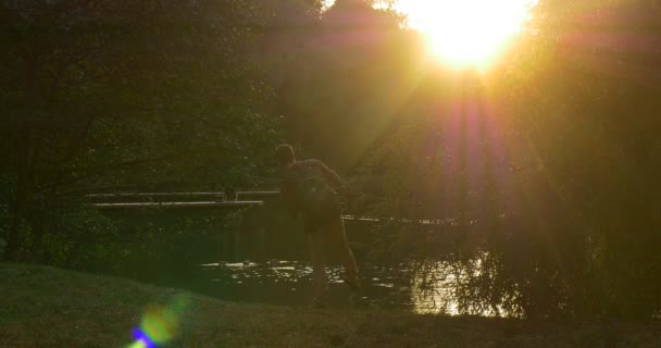 Человек с рюкзаком Мужской силуэт Солнце светит на камеру Прогулка по озеру Банк человек взял рюкзак и сидит Человек сидит и глядя на воду — стоковое видео