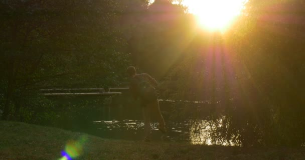 Uomo con lo zaino La silhouette dell'uomo Sole brilla davanti alla macchina fotografica Passeggiata da Lake Bank L'uomo ha tolto lo zaino e si siede L'uomo è seduto e guarda l'acqua — Video Stock