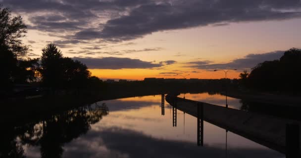 Presa en la presa del río en el dique de la ciudad apoya la reflexión del agua lisa casas residenciales siluetas de árboles de puesta de sol amarillo distante brillante — Vídeos de Stock