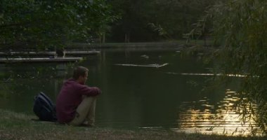 Man River Lake Bank Sırt Çantası oturuyor Yakın Man Su Duck içine Gıda atar Su Sun Feflection Yüzey İzi ile Yüzme olduğunu
