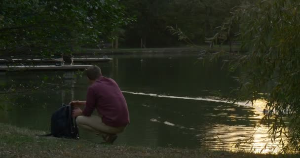 Man is Sitting at The River Lake Bank Took His Backpack Procurando algo na mochila Wild Duck Mallard is Swimming Trace on the Water Surface — Vídeo de Stock