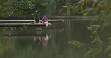 Sırt Çantası ile Man Lake River Şezlong Adam Ahşap Pier comes Off Onun Sırt Çantası Kapalı Pier Katta Oturur Bacakları Aşağı Su Ducks için